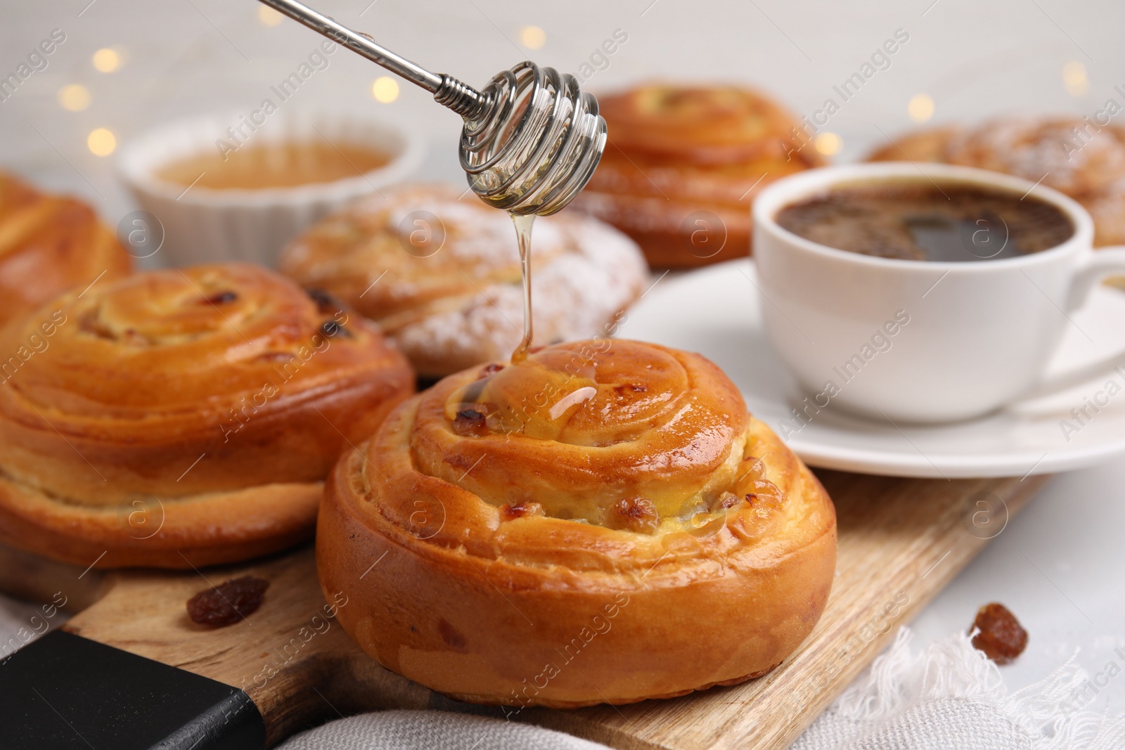 Photo of Pouring honey onto delicious rolls with raisins at table, closeup. Sweet buns