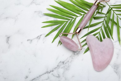Gua sha stone, face roller and green leaves on white marble table, flat lay. Space for text