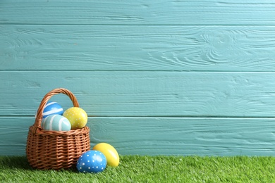 Wicker basket with painted Easter eggs on green grass against wooden background, space for text