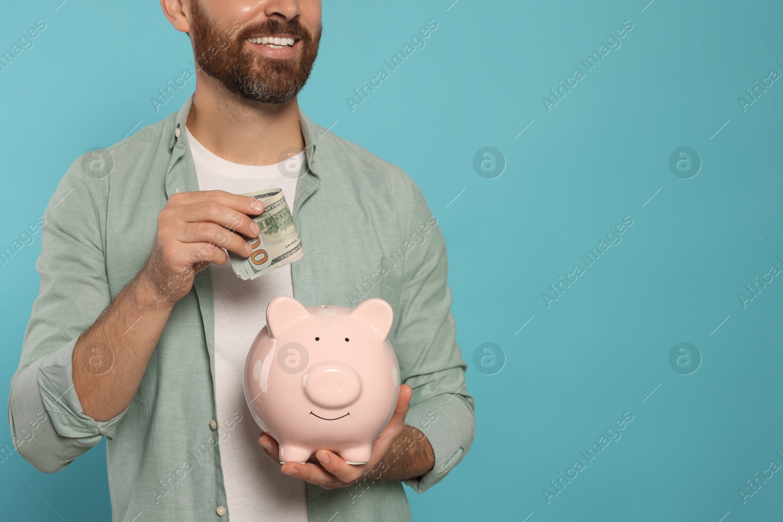 Photo of Happy man putting money into piggy bank on light blue background, closeup. Space for text