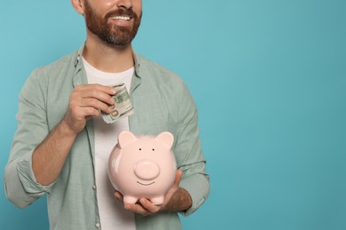 Photo of Happy man putting money into piggy bank on light blue background, closeup. Space for text