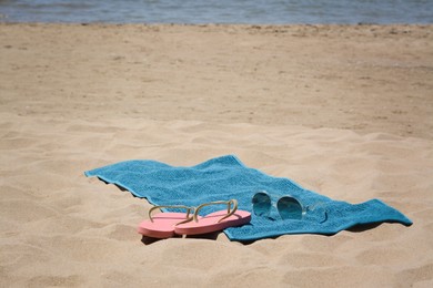 Photo of Soft blue beach towel, pink flip flops and sunglasses on sand near sea. Space for text