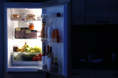 Photo of Open refrigerator full of products in kitchen at night