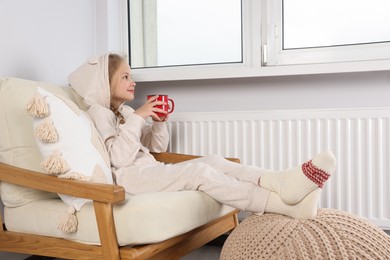 Little girl with cup of hot drink near heating radiator indoors