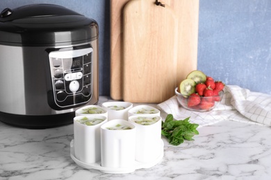 Cups of homemade fruit yogurt near multi cooker on marble table