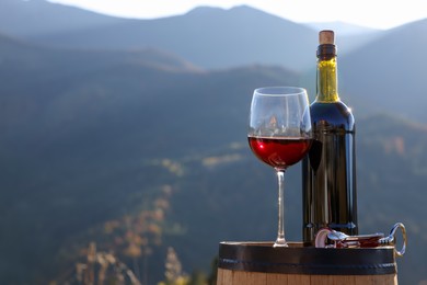 Photo of Corkscrew, glass and bottle of red wine on barrel against mountain landscape, space for text