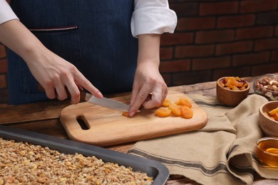 Photo of Making granola. Woman cutting dry apricots at wooden table, closeup