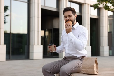 Photo of Lunch time. Businessman eating hamburger and holding paper cup of coffee on bench outdoors