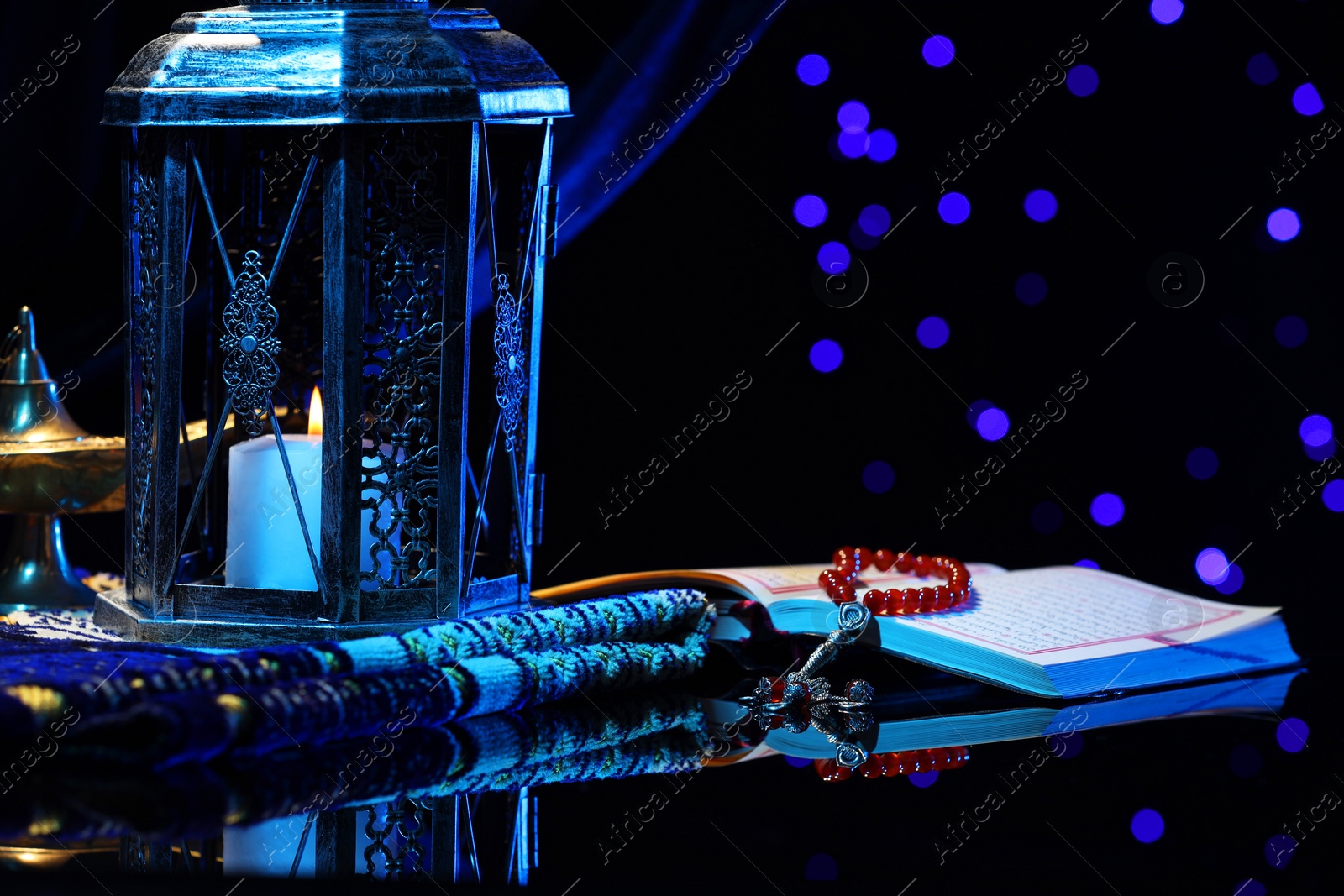 Photo of Arabic lantern, Quran, misbaha, Aladdin magic lamp and folded prayer mat on mirror surface against blurred lights at night. Space for text