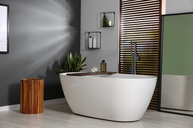 Stylish bathroom interior with ceramic tub, candles and care products on wooden bath tray