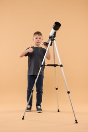 Photo of Cute little boy with telescope showing thumb up on beige background
