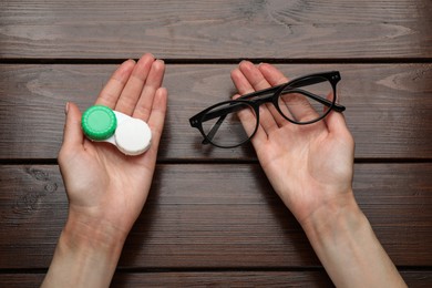 Woman holding case with contact lenses and glasses at wooden table, top view