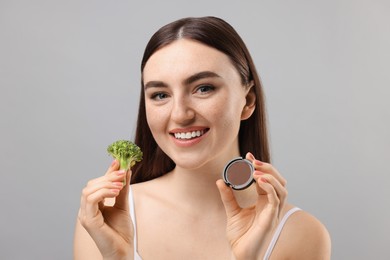 Photo of Smiling woman making fake freckles with broccoli and cosmetic product on grey background