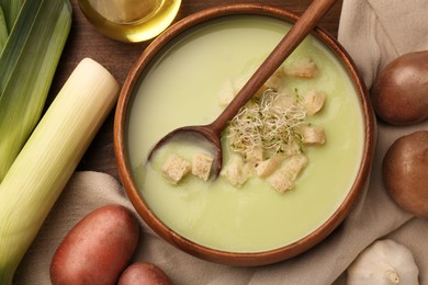 Bowl of tasty leek soup, spoon and ingredients on wooden table, flat lay