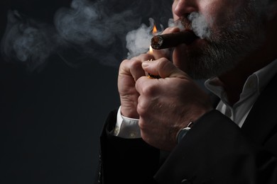 Bearded man lighting cigar on dark grey background, closeup. Space for text