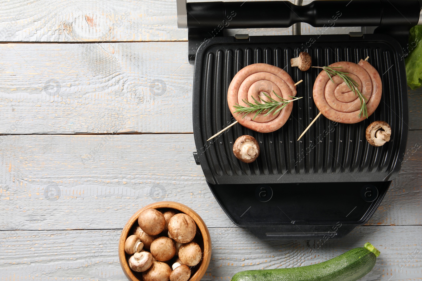 Photo of Electric grill with homemade sausages, rosemary and vegetables on rustic wooden table, flat lay. Space for text