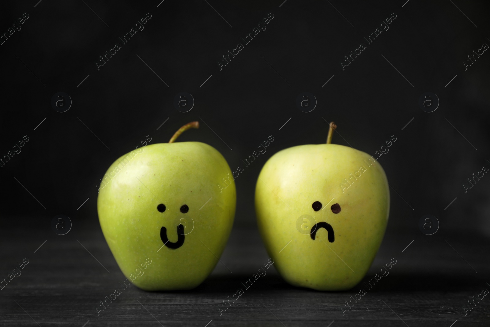 Photo of Apples with drawings of sad and happy faces on table against dark background. Depression symptoms