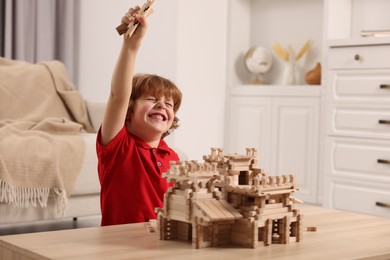 Photo of Emotional boy playing with wooden castle at table in room. Child's toy