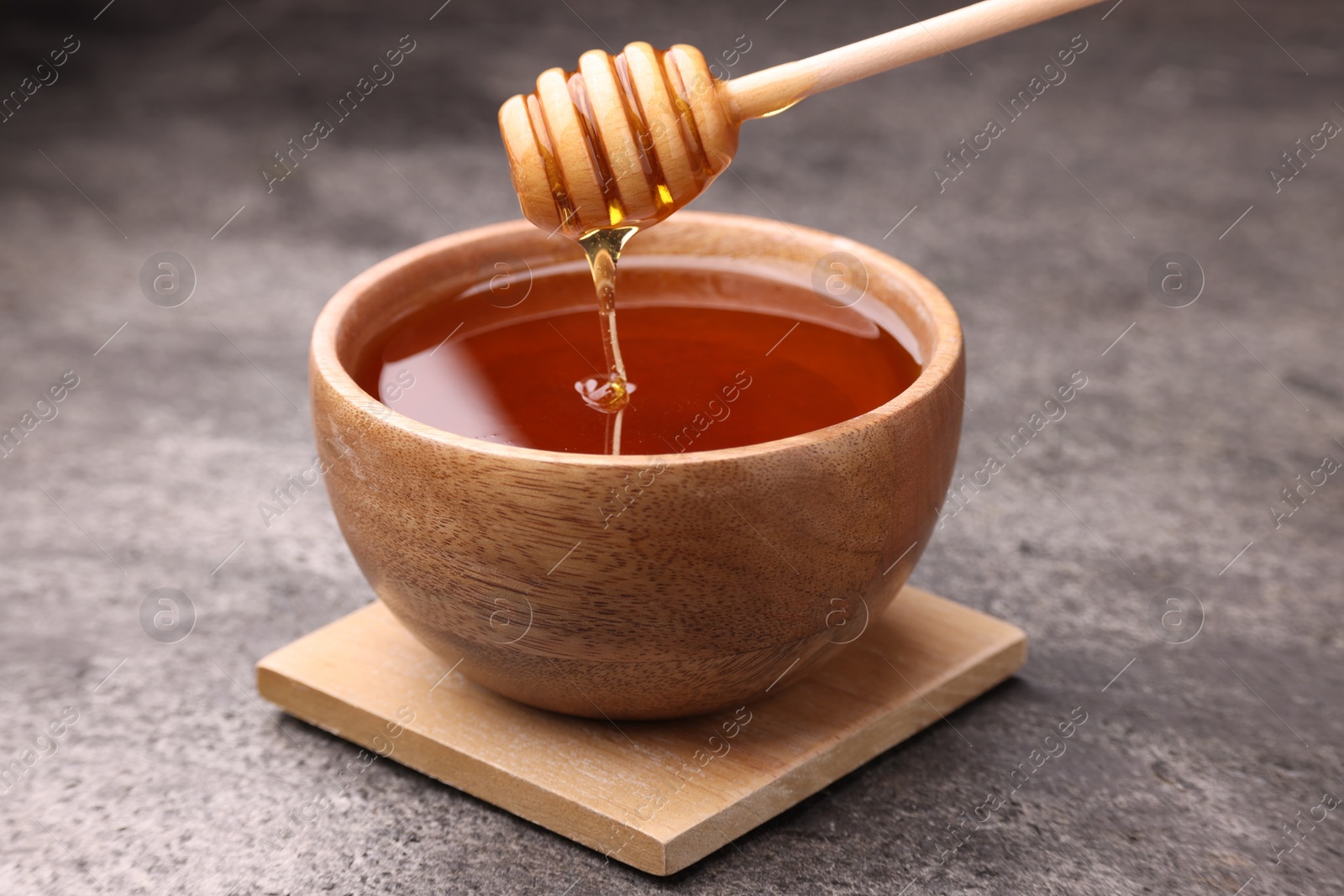 Photo of Pouring delicious honey from dipper into bowl on grey textured table, closeup