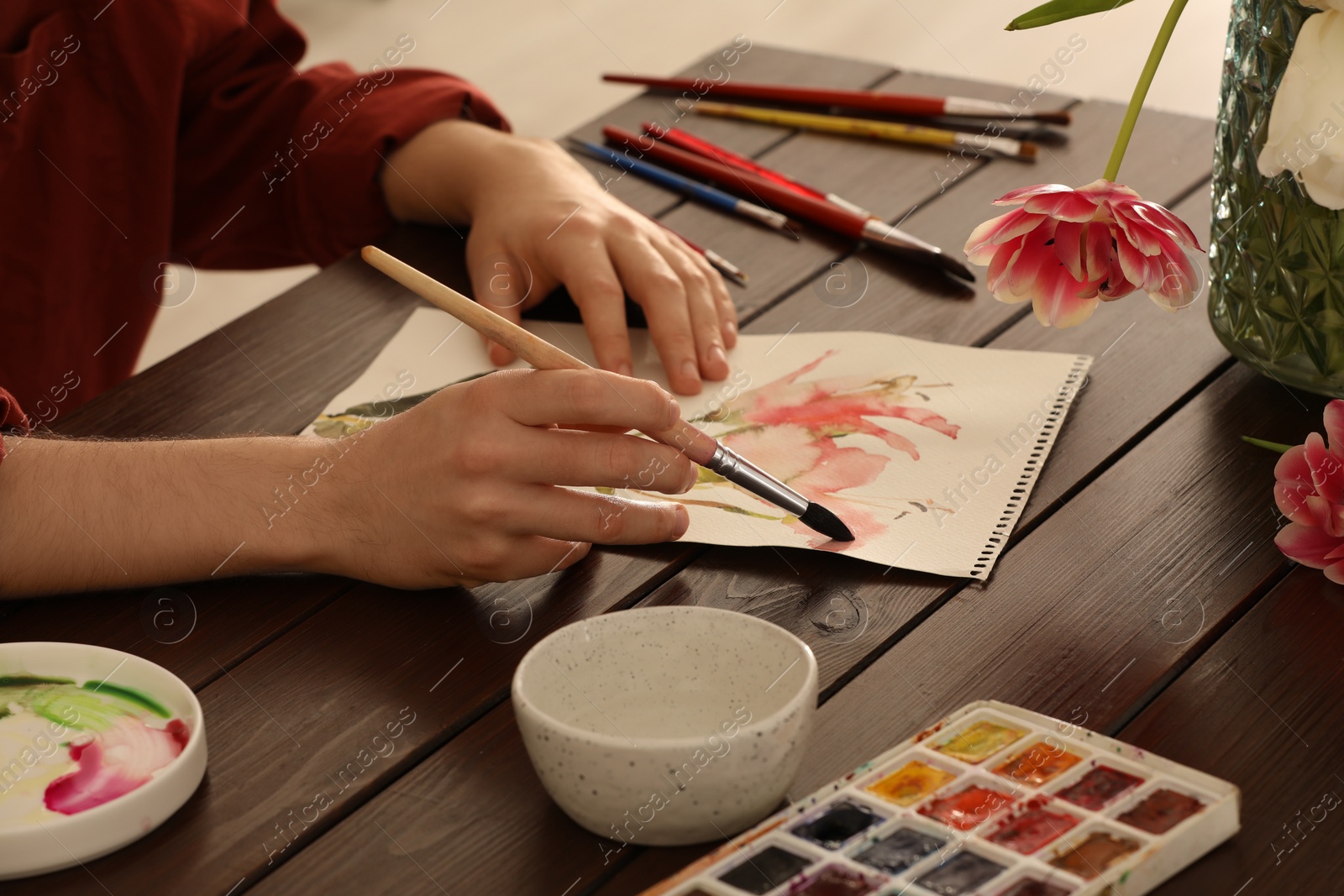 Photo of Woman painting flowers with watercolor at wooden table, closeup. Creative artwork