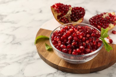 Photo of Ripe juicy pomegranate grains in bowl and green leaves on white marble table, space for text