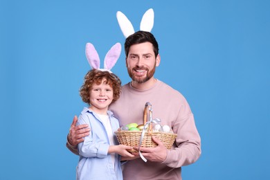 Photo of Happy father and son in cute bunny ears headbands holding Easter basket with painted eggs on light blue background