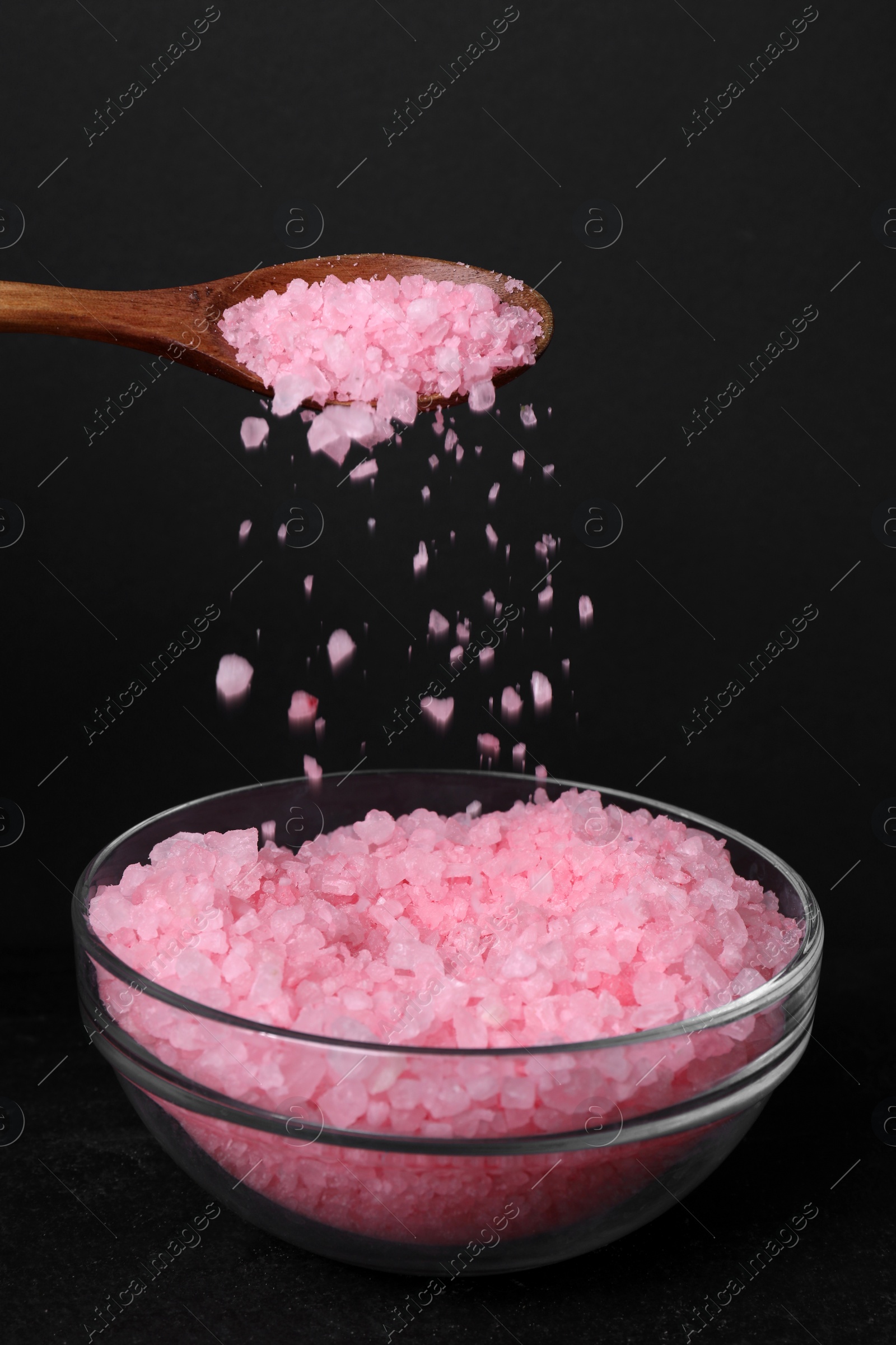 Photo of Pouring pink sea salt from wooden spoon into glass bowl against black background, closeup