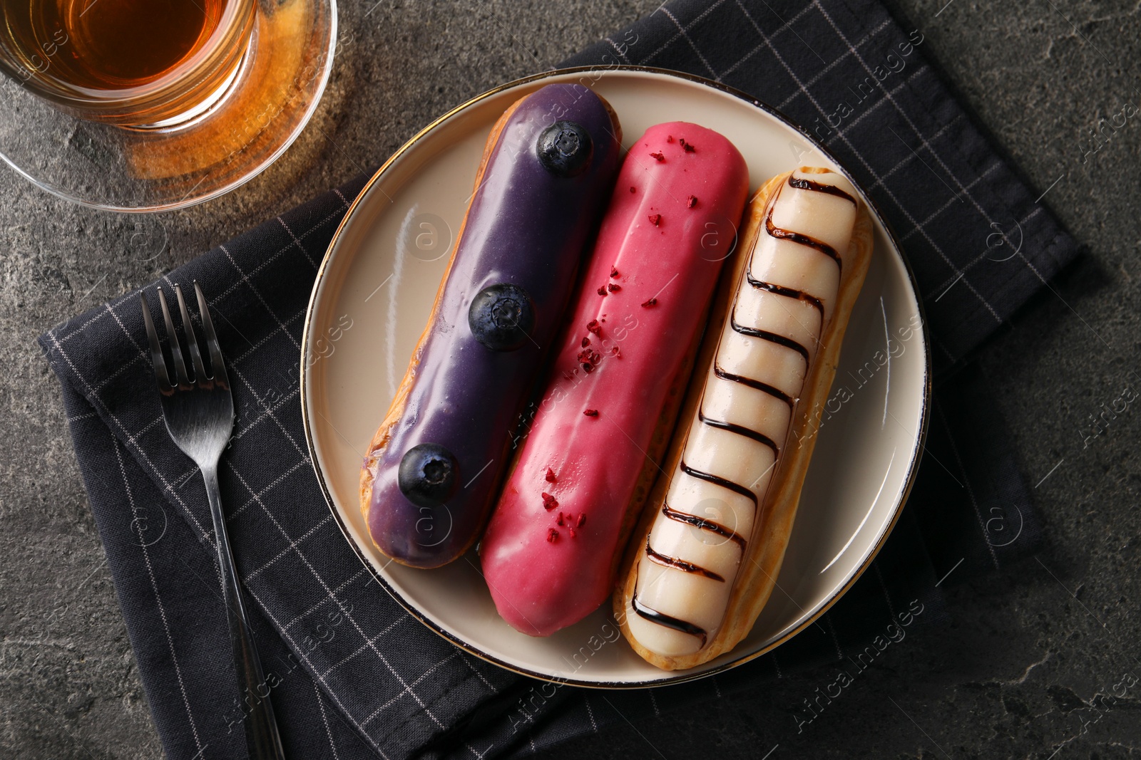 Photo of Different tasty glazed eclairs served with tea on grey textured table, flat lay