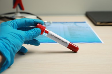 Photo of Laboratory worker holding tube with blood sample and label Liver Function Test at wooden table, closeup
