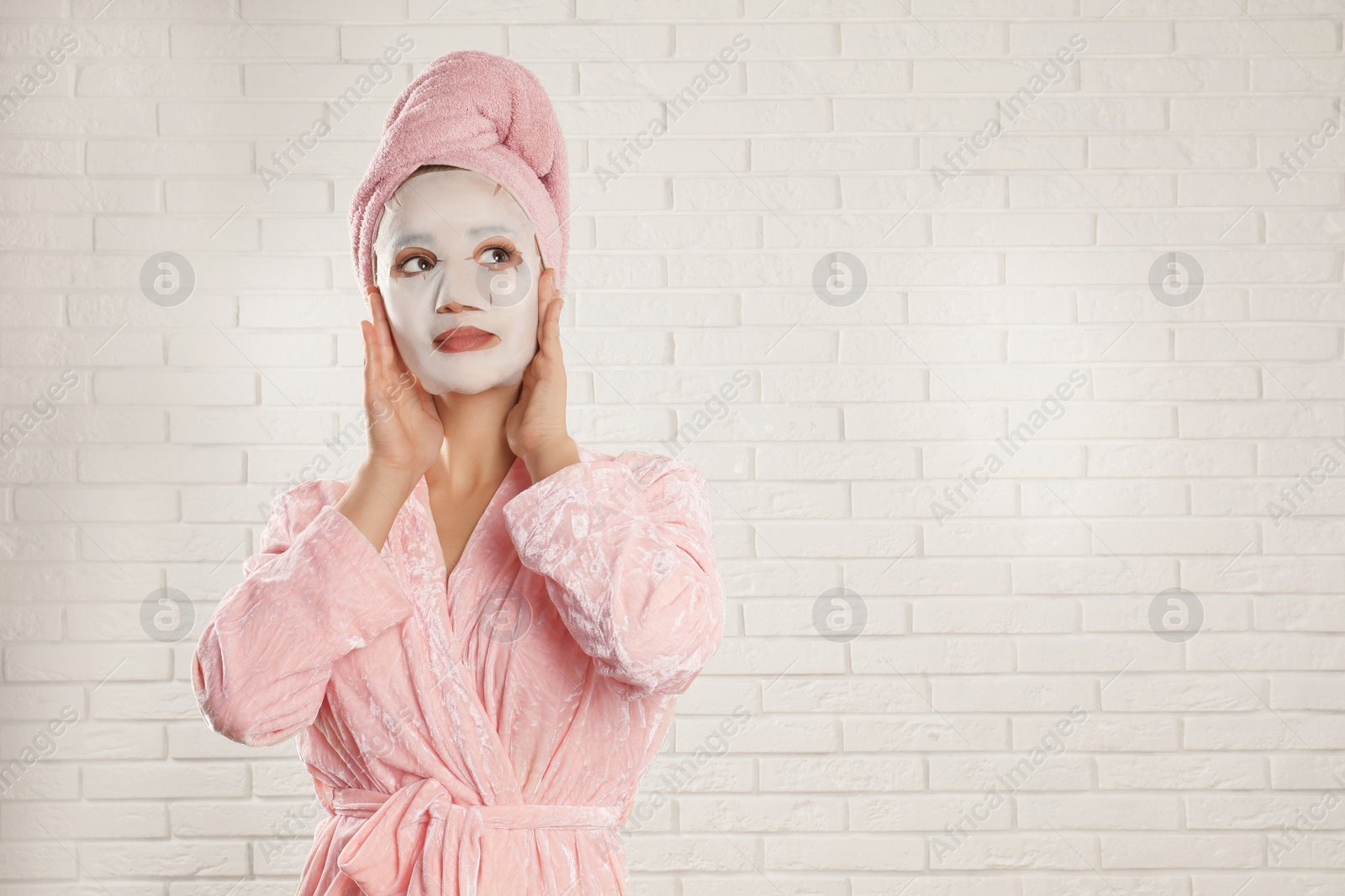 Photo of Young woman in bathrobe with cotton facial mask near white brick wall. Space for text