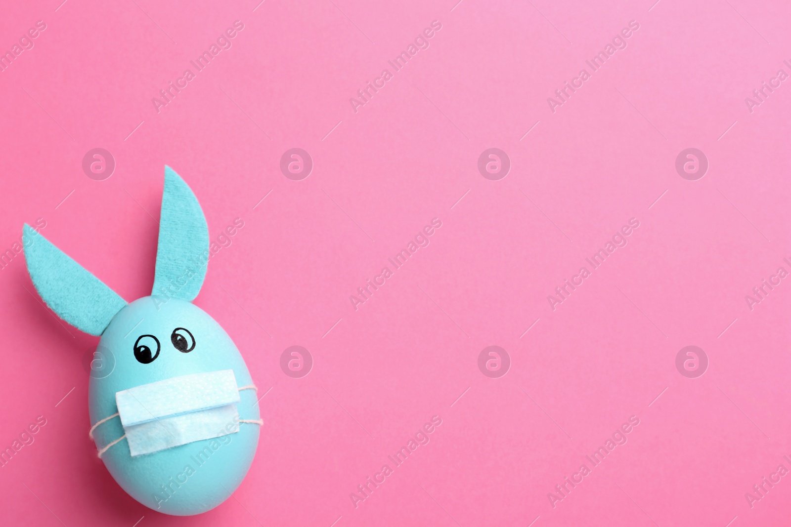 Photo of Painted egg decorated with bunny ears, protective mask and space for text on pink background, top view. Easter holiday during COVID-19 quarantine