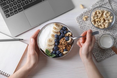 Photo of Woman eating tasty granola at white wooden table with laptop, top view