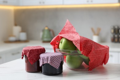 Photo of Jars of jams and apples in bowl covered with beeswax food wrap on white table indoors