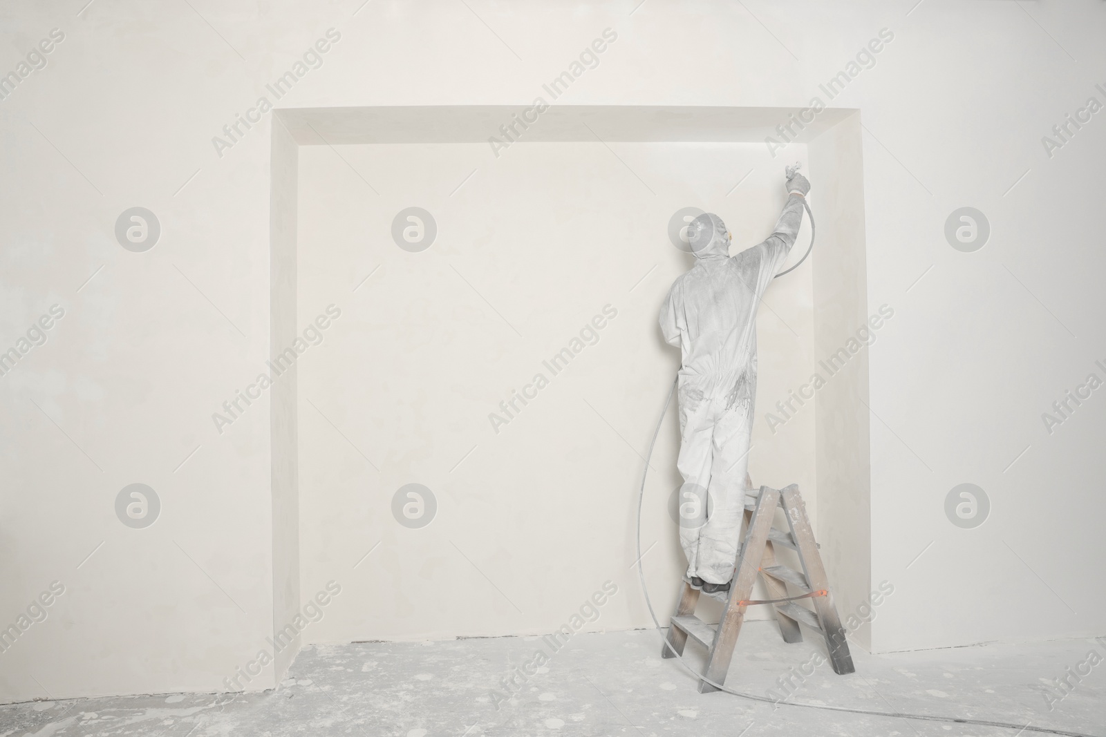 Photo of Decorator in protective overalls painting wall with spray gun indoors