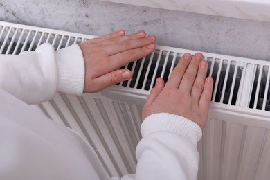 Photo of Girl warming hands on heating radiator indoors, closeup