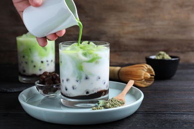 Photo of Person pouring green matcha into glass with milk bubble tea at black wooden table, closeup