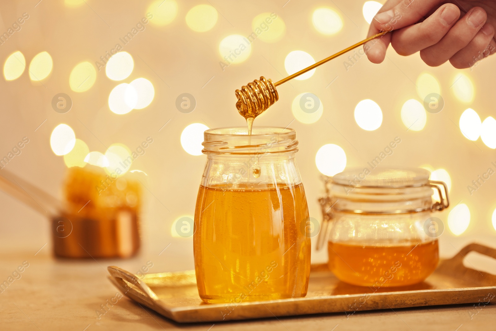 Photo of Woman holding dipper over jar with honey on table against blurred lights