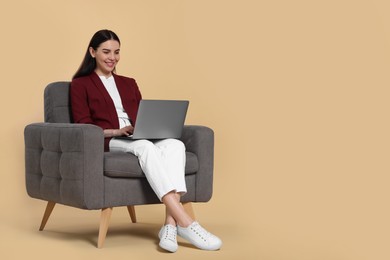 Photo of Happy woman with laptop sitting in armchair on beige background, space for text