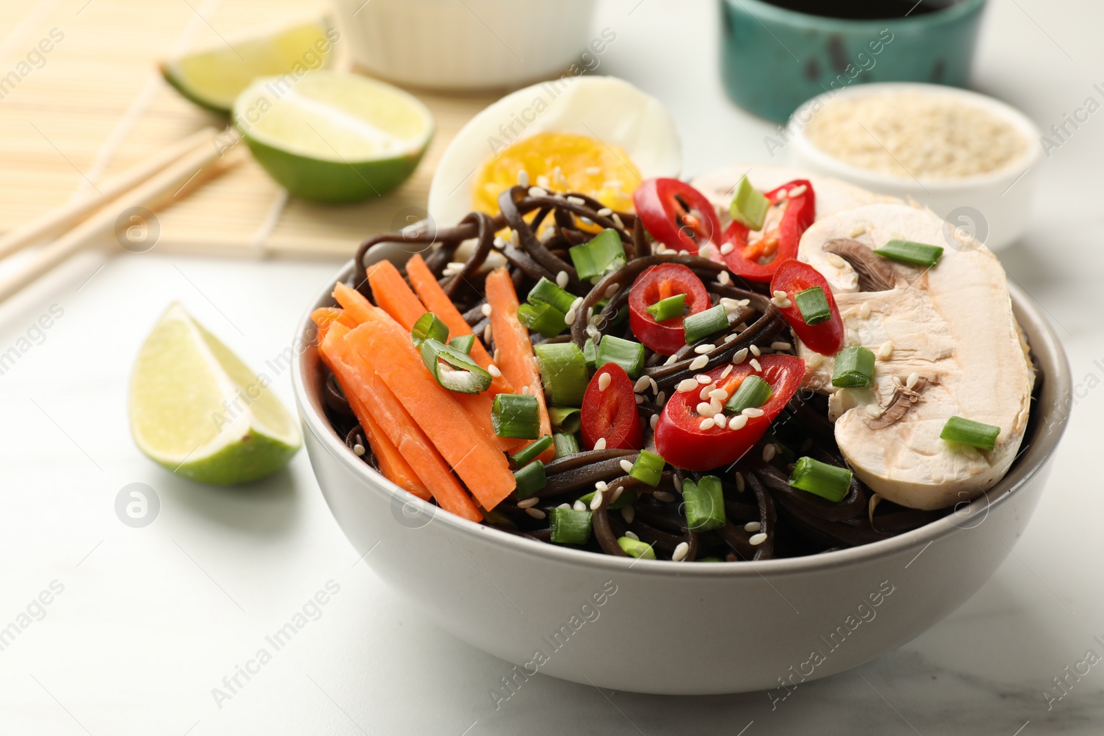 Photo of Tasty buckwheat noodles (soba) with chili pepper, egg, carrot and mushrooms in bowl on white marble table, closeup