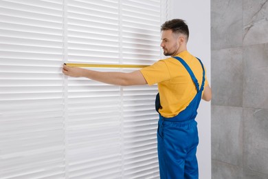 Photo of Worker in uniform using measuring tape while installing horizontal window blinds indoors