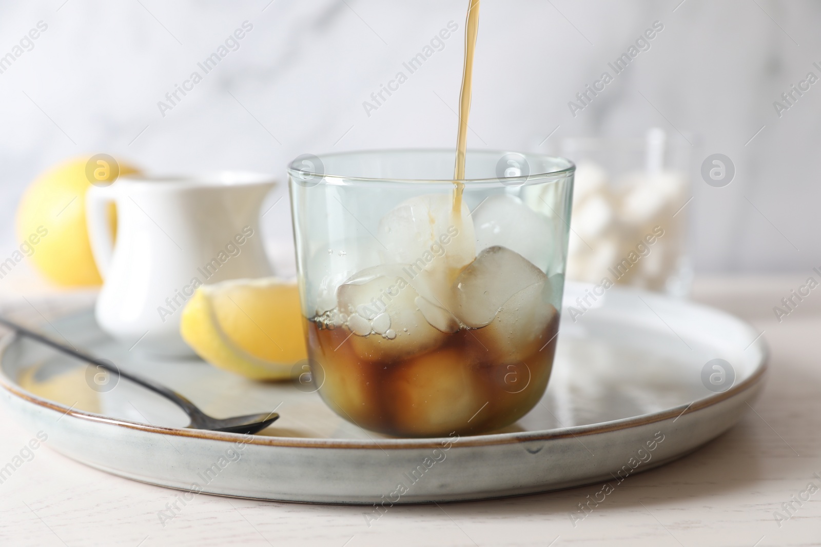 Photo of Pouring coffee into glass with ice cubes at white wooden table, closeup