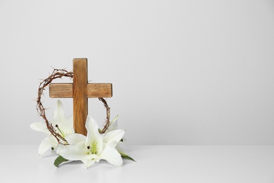 Photo of Wooden cross, crown of thorns and blossom lilies on table against light background, space for text