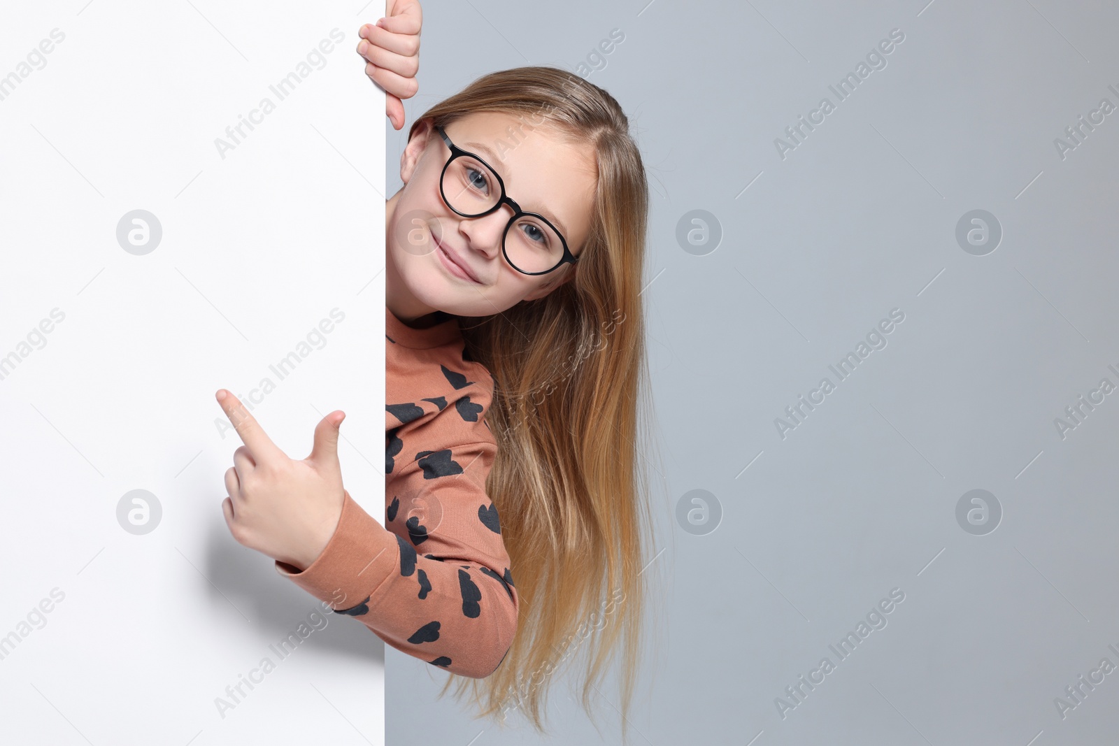 Photo of Cute girl looking out of placard and pointing on it against light grey background. Space for text