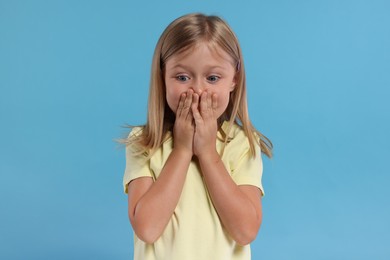 Embarrassed little girl covering mouth with hands on light blue background