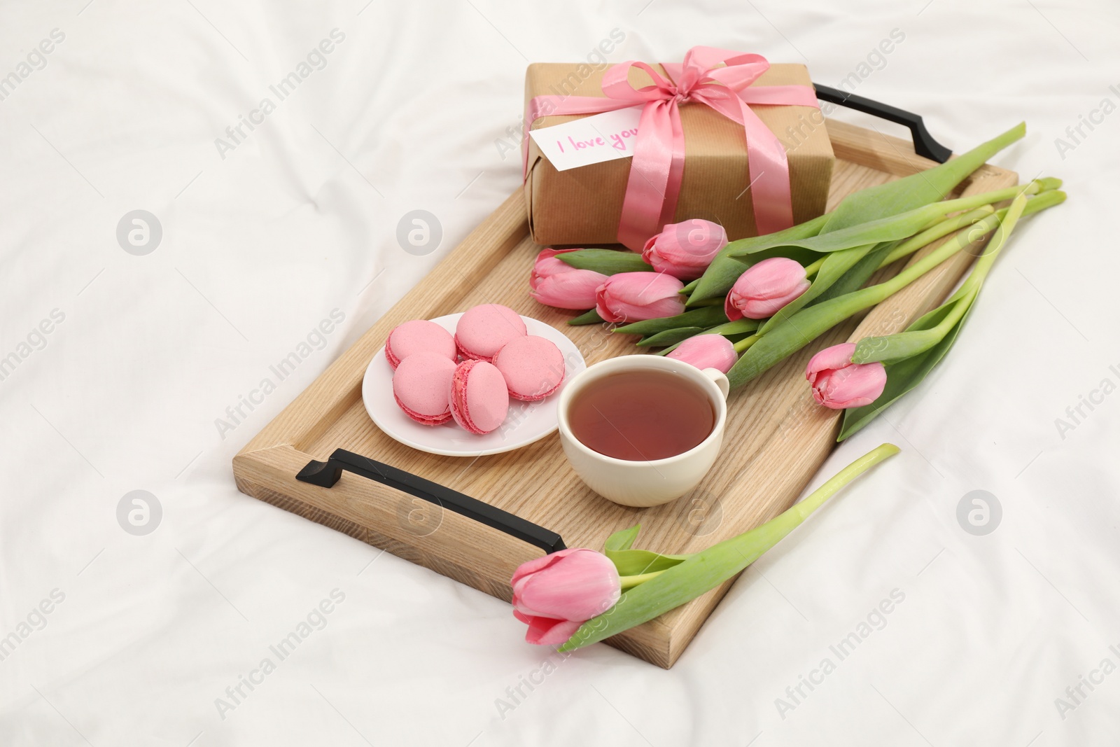 Photo of Tasty breakfast served in bed. Delicious macarons, tea, flowers, gift box and card with phrase I Love You on tray