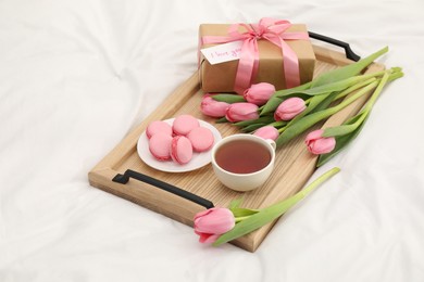 Photo of Tasty breakfast served in bed. Delicious macarons, tea, flowers, gift box and card with phrase I Love You on tray