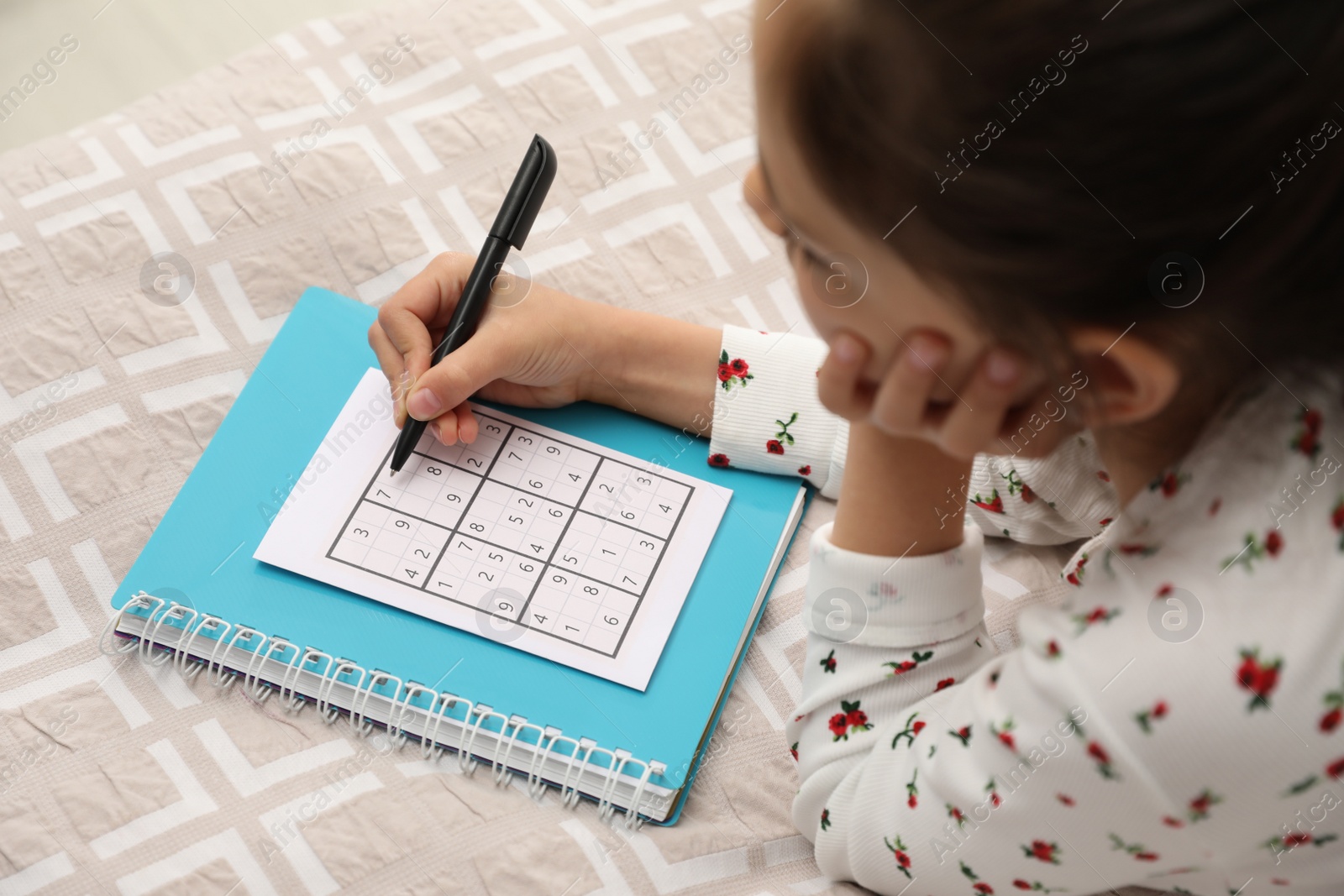 Photo of Little girl solving sudoku puzzle on bed