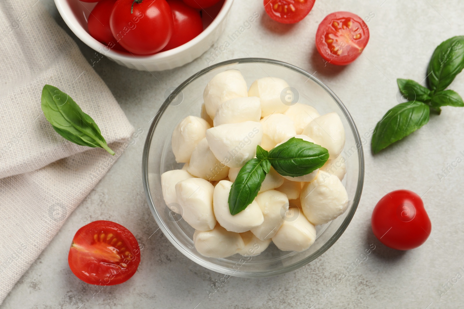 Photo of Delicious mozzarella balls in glass bowl, tomatoes and basil leaves on light gray table, flat lay
