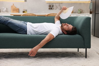 Photo of Man waving white hand fan to cool himself on sofa at home