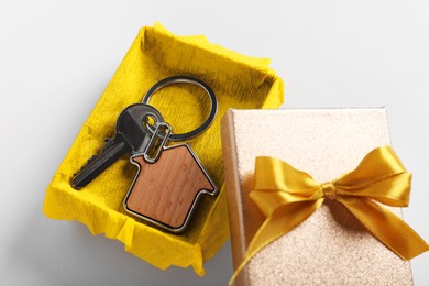 Key with trinket in shape of house and gift box on light grey background, closeup. Housewarming party